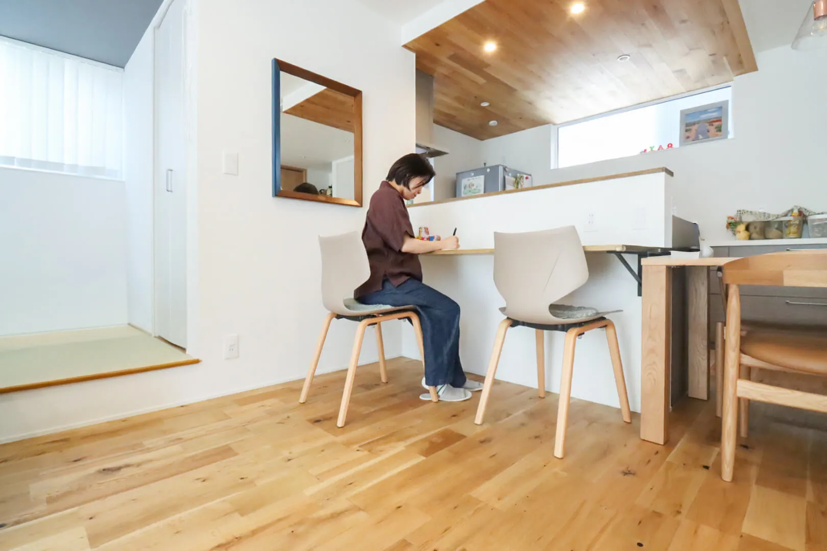 キッチンカウンター横の造作デスクで書き物をする女性。天井には木目パネル。
