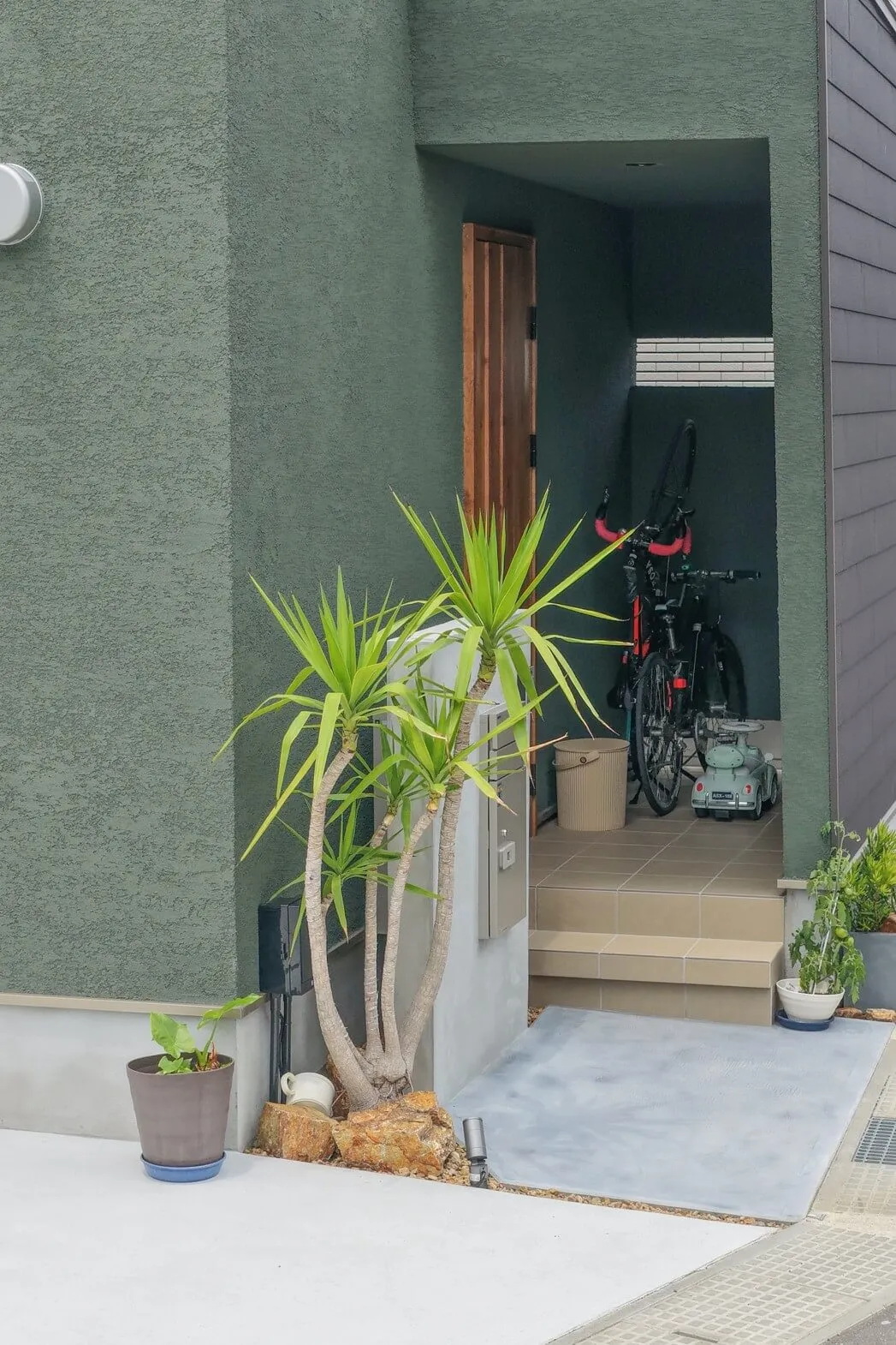 株式会社シンズクラフトによる注文住宅の玄関ポーチで、深みのあるグリーンの外壁に植栽が映え、自転車も置ける奥行きのある土間スペースを確保した、こだわり溢れる工務店の家づくり。