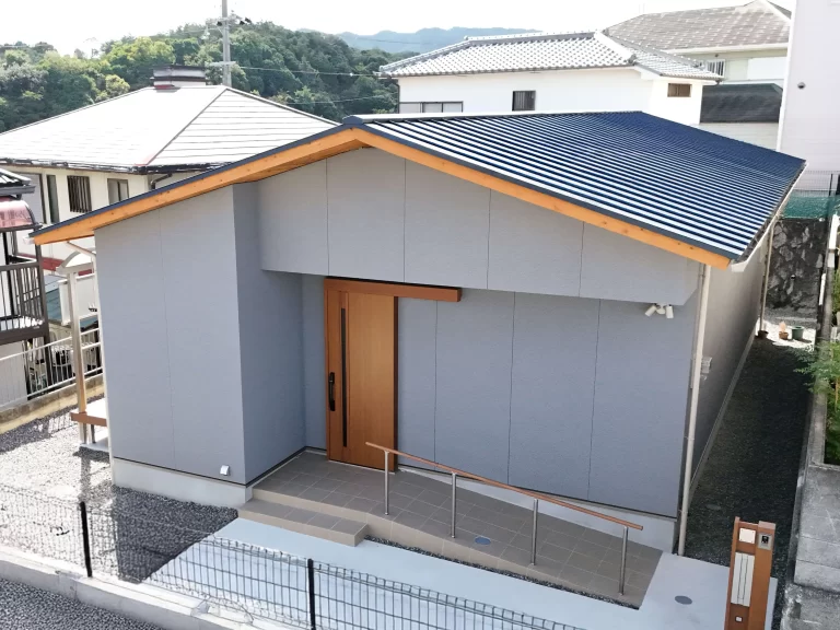 株式会社シンズクラフトによる平屋の注文住宅の外観写真で、落ち着いたグレーの外壁に木目の玄関引き戸と軒天が映え、バリアフリーに配慮した手すり付きのスロープが設置された現代的な工務店の家づくり。