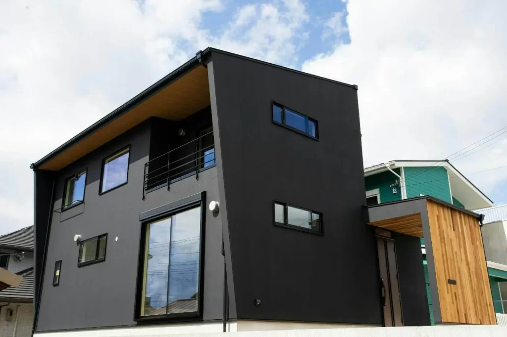 青い空と雲を背景に、黒いガルバリウム鋼板の外壁と木目調の軒天がシャープでモダンな印象を与える、傾斜のあるデザインが特徴的な2階建ての注文住宅の外観写真。