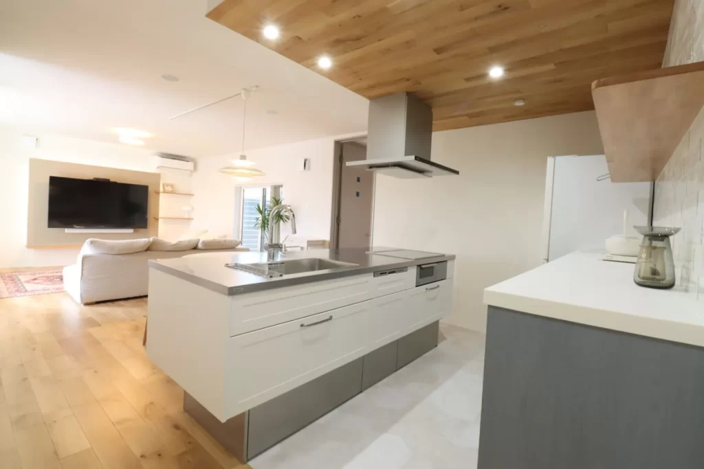 白を基調とした清潔感のあるアイランドキッチンと木目調の下がり天井が調和したLDKの内観写真で、リビング側には壁掛けテレビやソファがゆったりと配置され、注文住宅ならではのこだわりを感じさせる明るく開放的な空間デザインです。