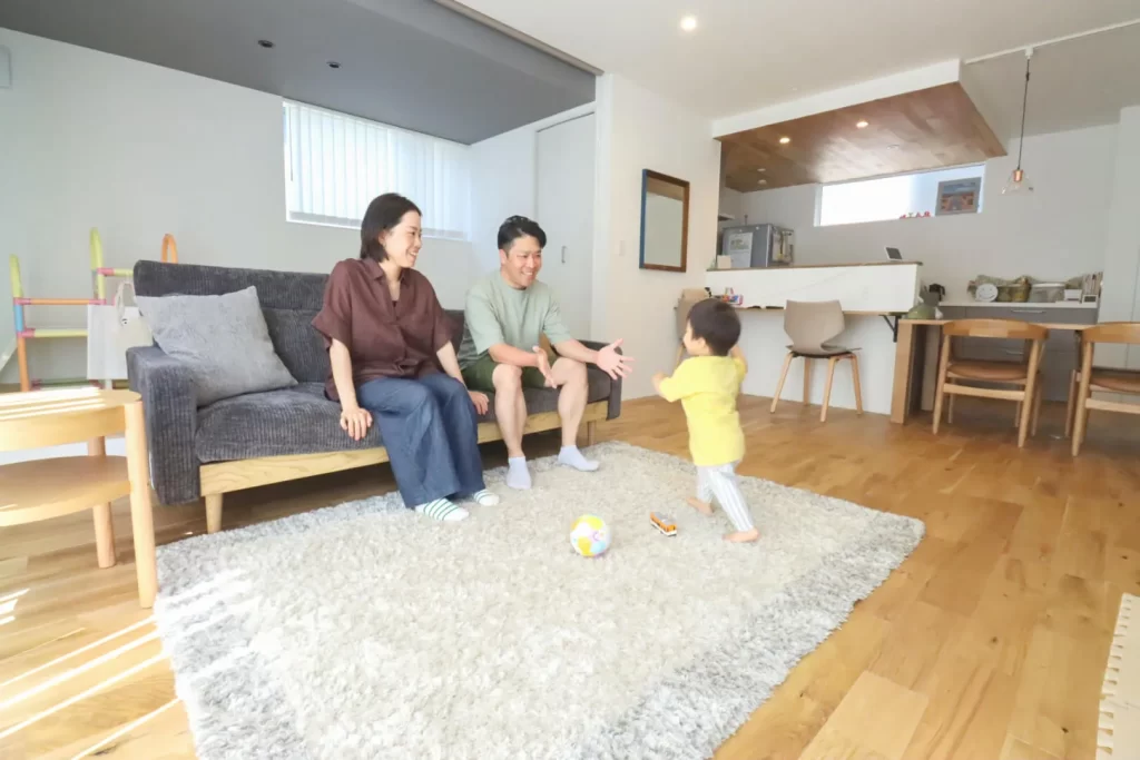 明るい採光が差し込むリビングで、ソファに座った夫婦が、広々としたラグの上でボールを追いかけている小さな子供を笑顔で見守っており、家族の暮らしを大切にした注文住宅のリビング空間の施工事例。