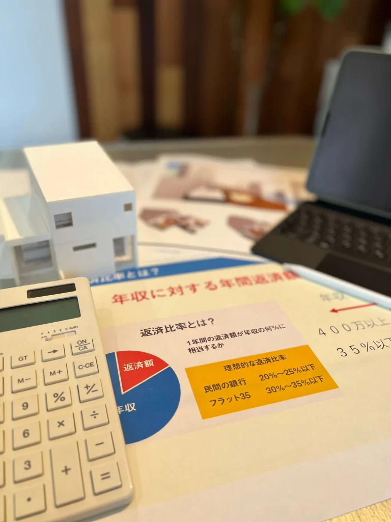 住宅模型と電卓、資料が並べられたデスクで、住宅ローンの年間返済額に対する年収の返済比率など、お客様の資金計画や予算に関する重要な情報を説明する資料に焦点を当てた写真。