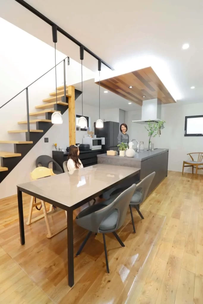 キッチンとダイニングテーブルを横並びに配置して家事動線を考慮したLDKの写真で、開放的な吹き抜け空間には黒い鉄骨手すりのスケルトン階段が設置され、家族が心地よく過ごせる注文住宅のモダンで機能的なインテリアデザインを示しています。
