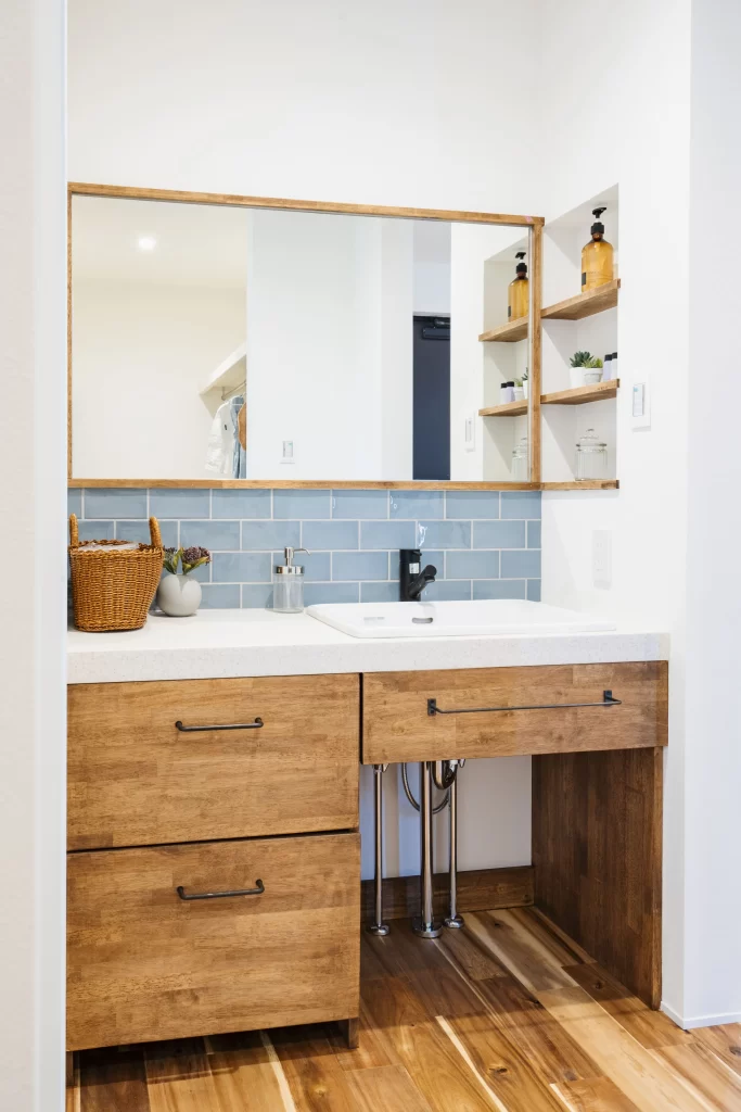 木の温もりを感じさせる引き出しや棚と爽やかな青いタイルを組み合わせたおしゃれな造作洗面台。壁面には使い勝手の良いニッチ収納や木枠の大きな鏡が設置され、注文住宅におけるオリジナリティあふれる水回り空間のデザイン事例です。