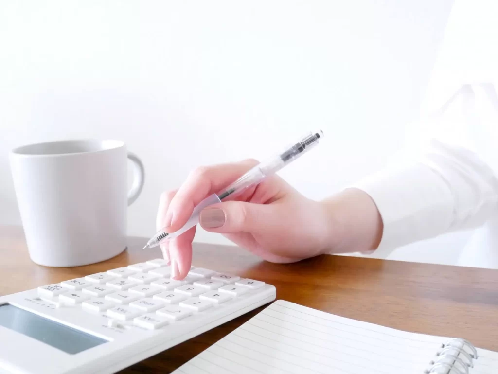 住宅購入に向けた予算立てやローン計算のイメージとして、白い電卓とノート、コーヒーカップが置かれた机で、女性がペンを持ちながら家づくりの資金計画を立てている手元の写真。