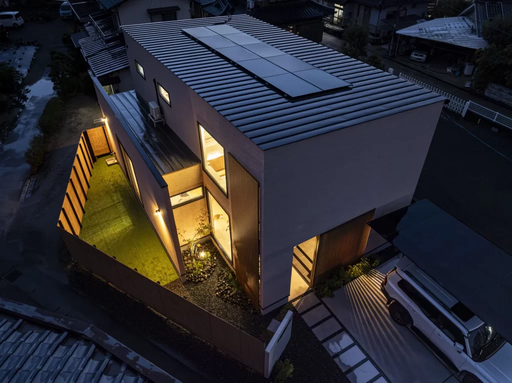 屋根にソーラーパネルを設置したスクエアデザインの注文住宅を夜間に上空から撮影した写真で、庭の照明や室内の灯りが美しく漏れる株式会社シンズクラフト設計の住まいの外観。