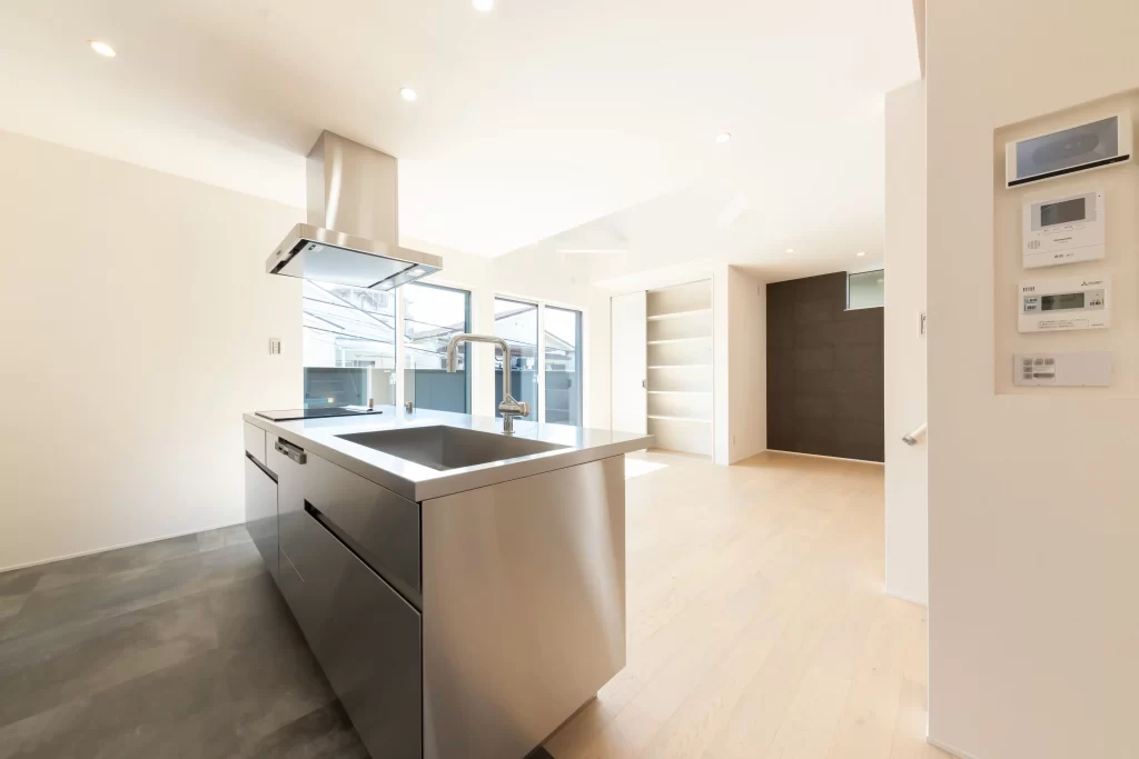 ステンレス製のアイランドキッチンを中心に据えた開放的なLDKで、白を基調とした壁と明るい無垢材のフローリングが特徴的な株式会社シンズクラフトによる注文住宅の施工事例。