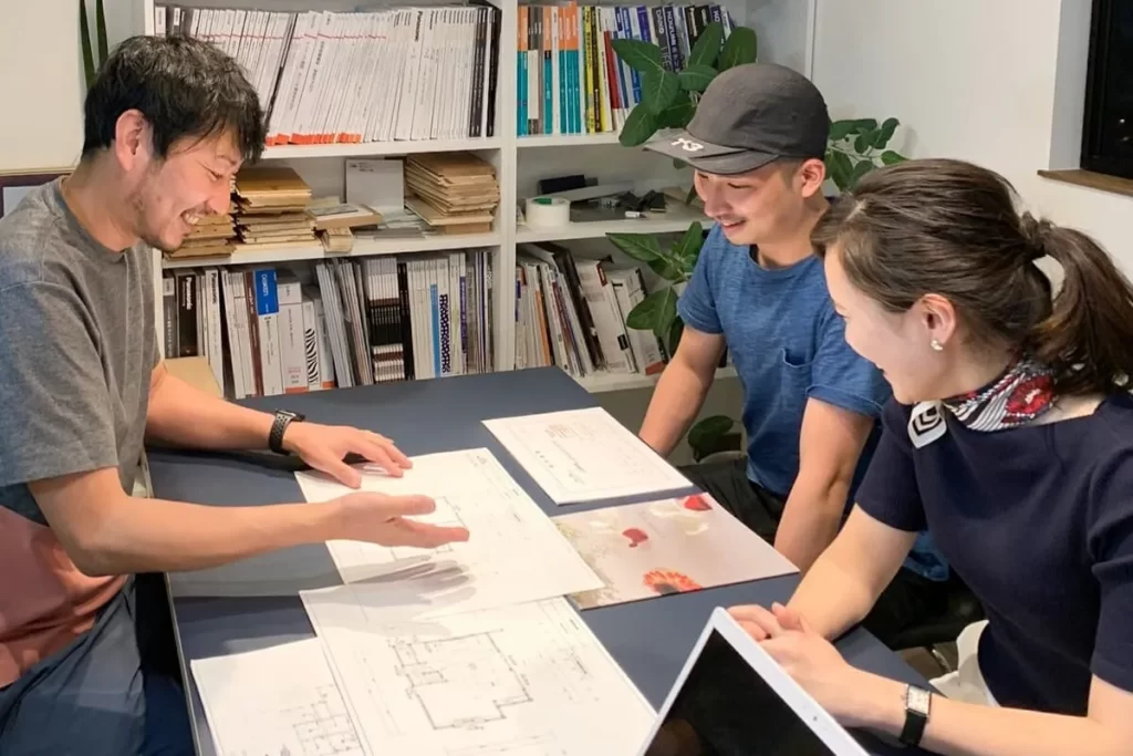 カタログやサンプルが並ぶ打ち合わせスペースで、図面を広げながらスタッフと施主夫婦が笑顔で家づくりの詳細プランを相談している株式会社シンズクラフトの打合せ風景。