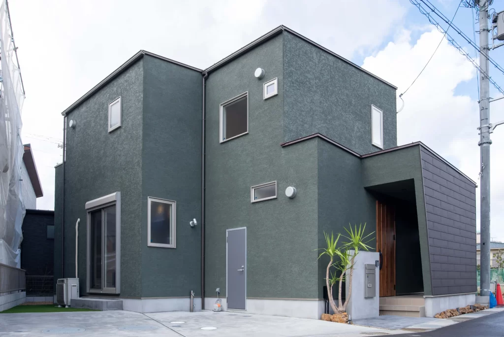 株式会社シンズクラフトが手掛けた注文住宅の施工事例で、深緑色の塗り壁が特徴的なモダンな外観デザインと、プライバシーに配慮した窓配置やスタイリッシュな玄関アプローチを備えた2階建て住宅。