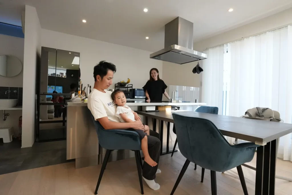 ステンレスのスタイリッシュなアイランドキッチンがあるLDKで、父親の膝の上に子供が座り、奥で母親が笑顔で見守っている株式会社シンズクラフトの施工事例における日常風景。