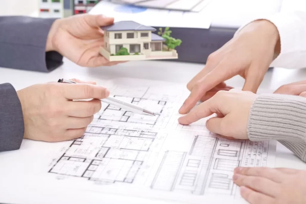 テーブルに広げられた住宅の間取り図を指差しながら、住宅模型を手に取って具体的な住まいのイメージを打ち合わせしている住宅会社と施主の手元の写真。