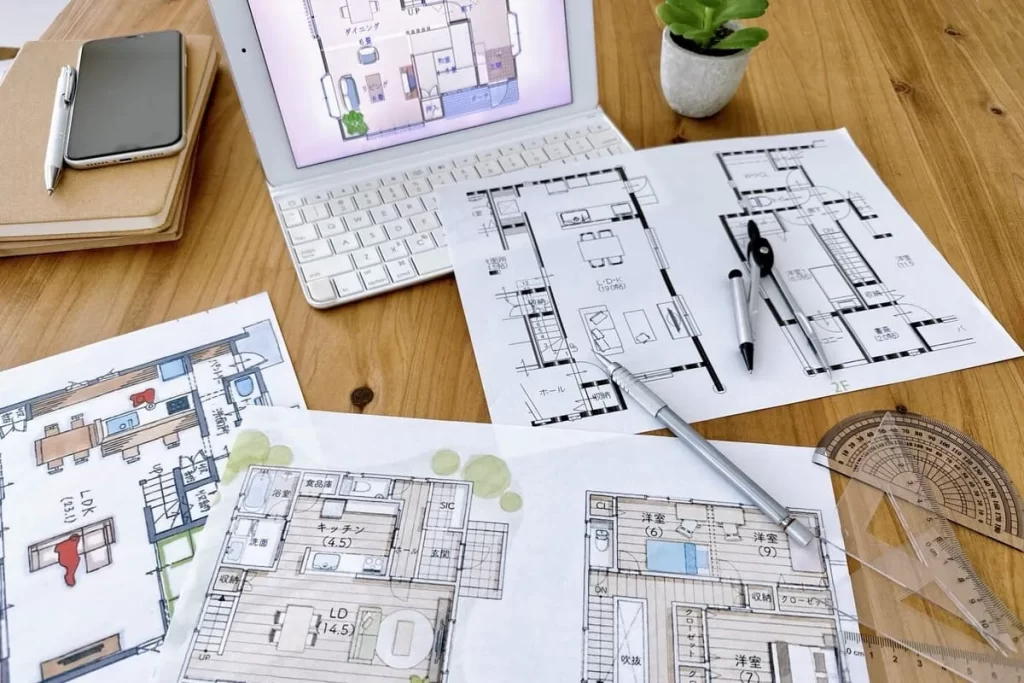 木目のテーブルの上に広げられた複数の間取り図、タブレット端末、コンパスや分度器などの設計道具が並び、自由設計による家づくりのプロセスを表現したイメージ写真。