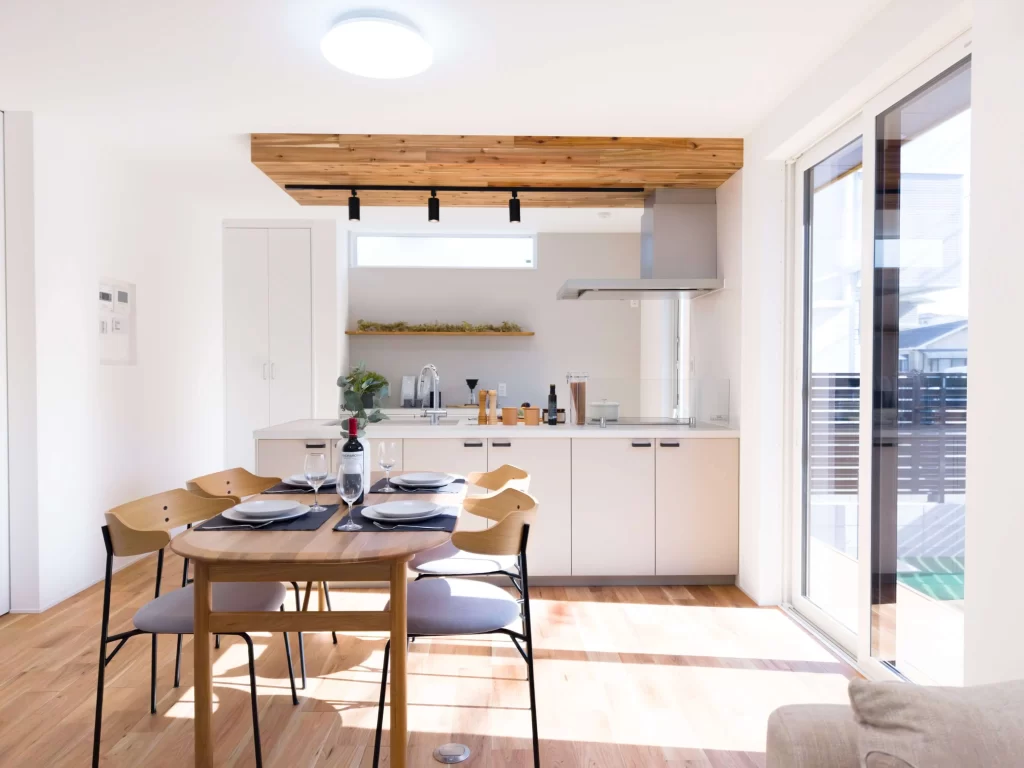 木の温もりを感じる下がり天井とライティングレールが印象的なキッチンから、自然光が差し込む明るいダイニングテーブルを望む開放的なLDKの内観写真。