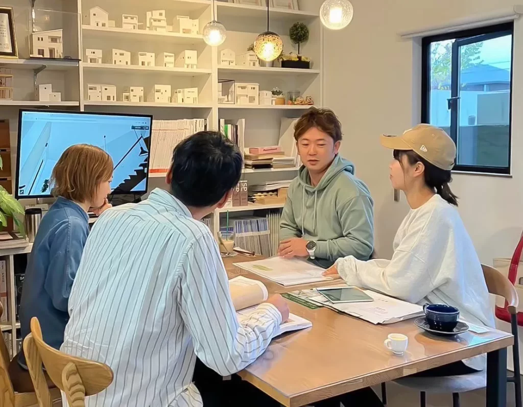 背景の棚に多くの住宅模型が並ぶ打ち合わせスペースで、モニターを見ながら家づくりの計画についてスタッフと施主が対面で相談している株式会社シンズクラフトの接客風景。