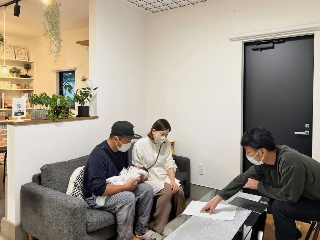 株式会社シンズクラフトの事務所にて、赤ちゃんを連れたご家族がスタッフとソファに座りながら、資料を基に家づくりや資金計画について真剣に相談を行っている打ち合わせの様子。