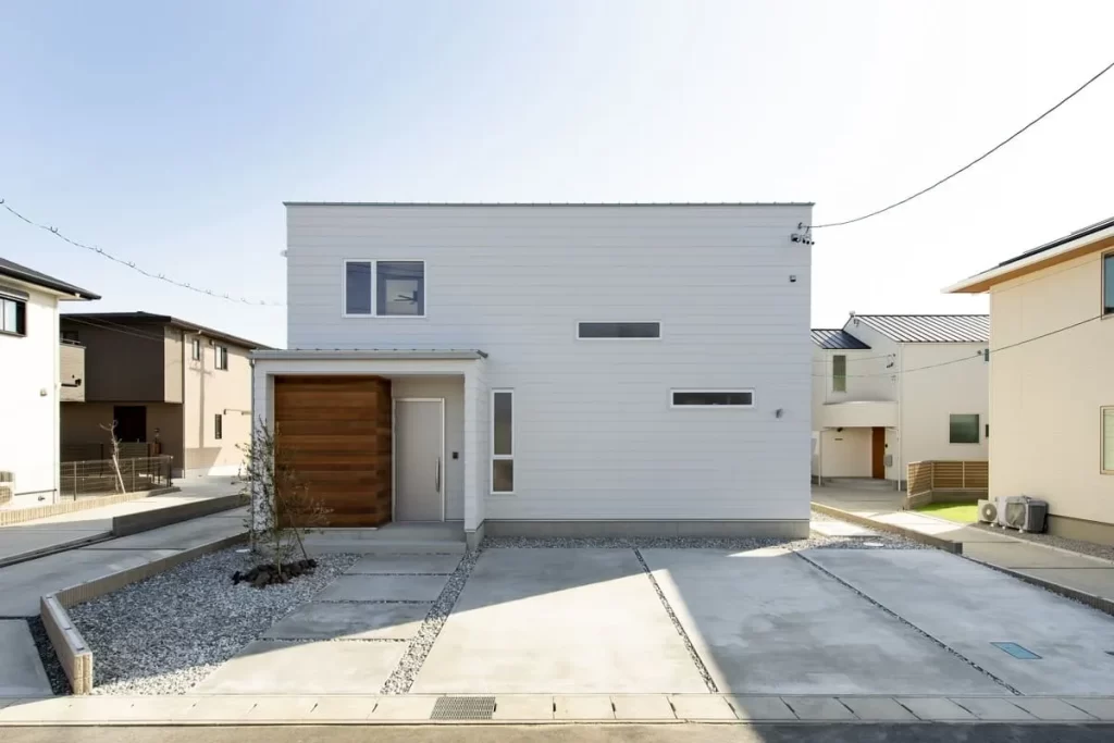 白のガルバリウム外壁に木目調の玄関ポーチがアクセントとなった、シンプルで洗練されたボックス型のフォルムが特徴的な株式会社シンズクラフトの注文住宅の外観写真。