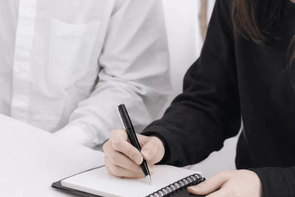 白いシャツを着た人物の隣で、黒い服を着た人物がリングノートにペンでメモを取っている様子を写した、打ち合わせやヒアリングを想起させるイメージ写真。