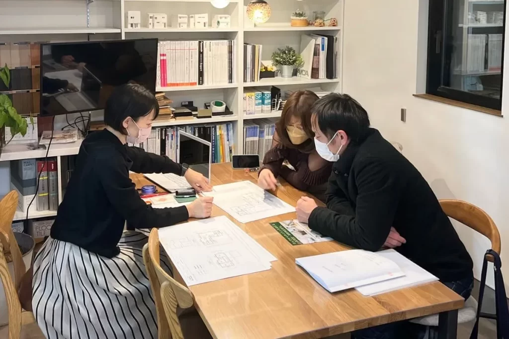 住宅模型やカタログが並ぶ打ち合わせスペースで、図面を広げながらスタッフと施主が家づくりの詳細について相談を行っている株式会社シンズクラフトでの打ち合わせ風景。