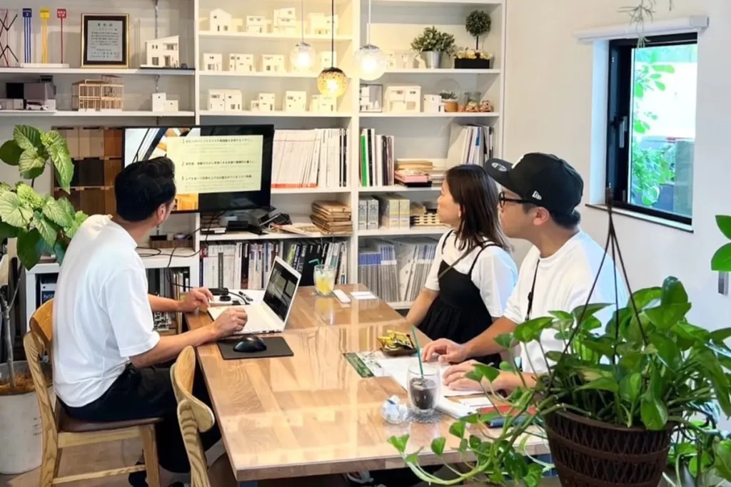 住宅模型が並ぶ棚を背景に、モニターに映し出された資料を見ながらスタッフと施主が詳細な設計プランについて相談を行っている株式会社シンズクラフトの打ち合わせ風景。