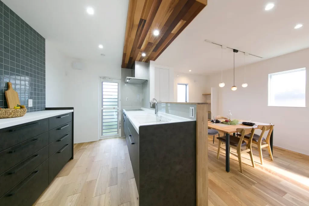 黒の対面キッチンと木目調の下がり天井がスタイリッシュな空間を演出し、奥にダイニングセットが配置された、光溢れる株式会社シンズクラフトの注文住宅の内観施工事例。