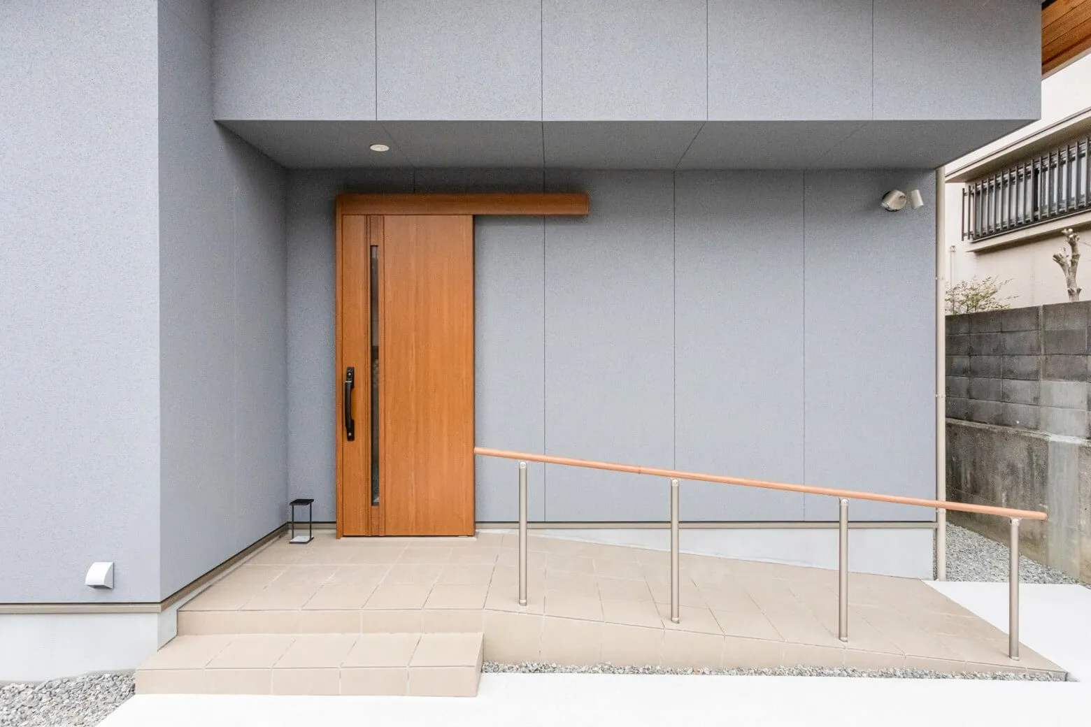 株式会社シンズクラフトが設計した注文住宅の玄関アプローチで、モダンなグレーの外壁に木目調の引き戸が映え、車椅子やベビーカーでも安心して移動できる手すり付きのスロープを完備した、バリアフリーな家づくりの施工事例。