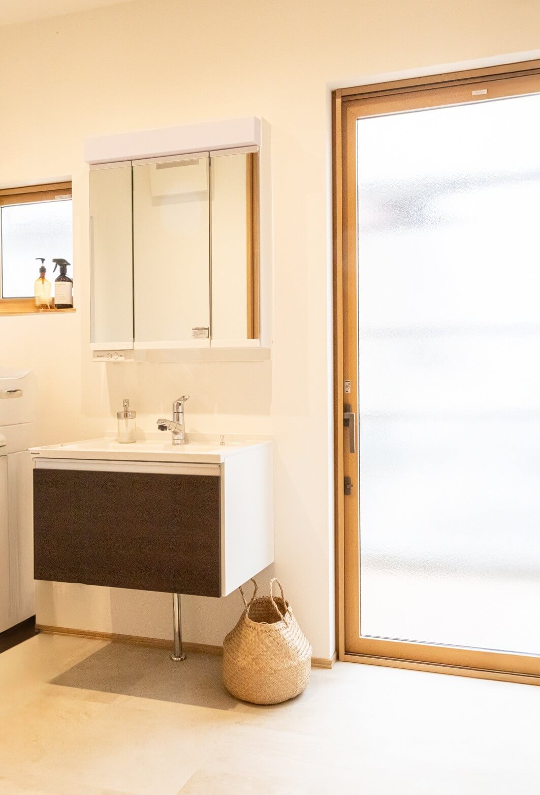 株式会社シンズクラフトが手掛けた注文住宅の洗面脱衣所で、ダークブラウンの洗面台と三面鏡、自然光が差し込むテラスドア、ナチュラルな編み込みバスケットが置かれた清潔感のある家づくりの施工事例。