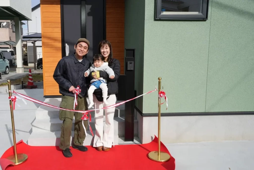 完成した新しい家の前で、笑顔でテープカットを行う若い夫婦と小さなお子様の家族写真。