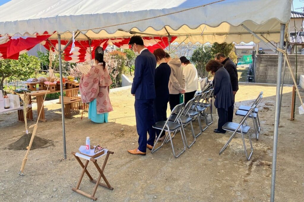 青空の下、テント内で神主が祝詞を奏上し、施主家族が参列している地鎮祭の風景。