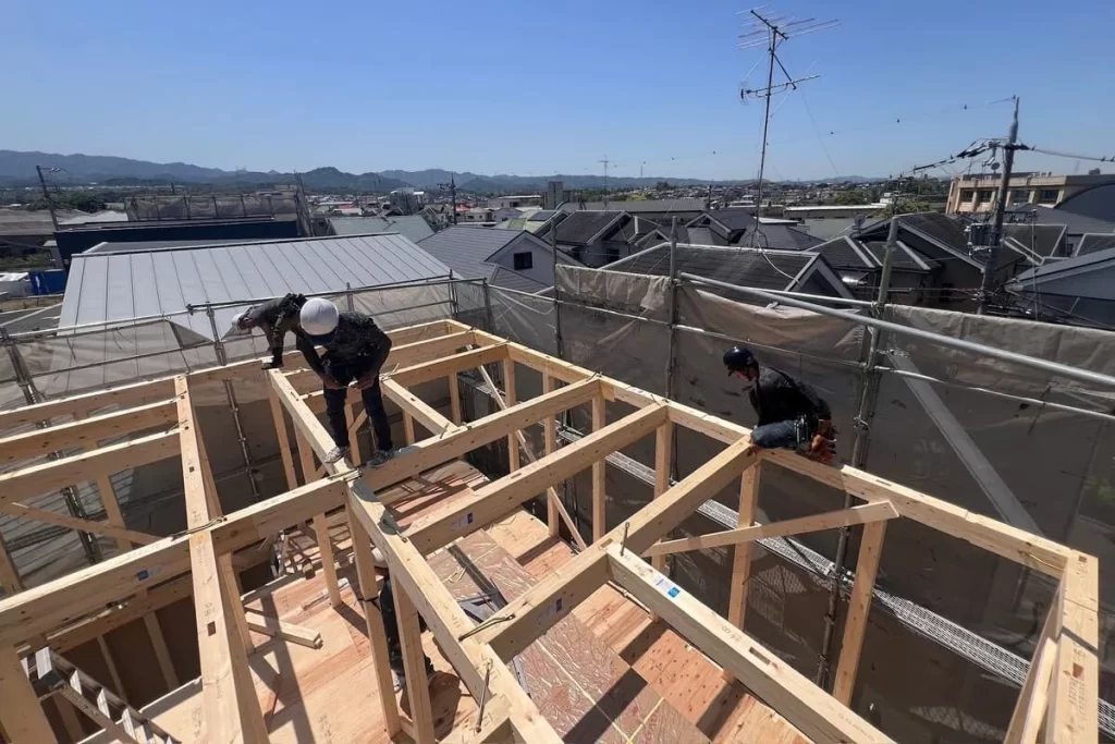 青空の下、住宅の木造骨組み(上棟)の上で作業を行う熟練の職人たちの様子。