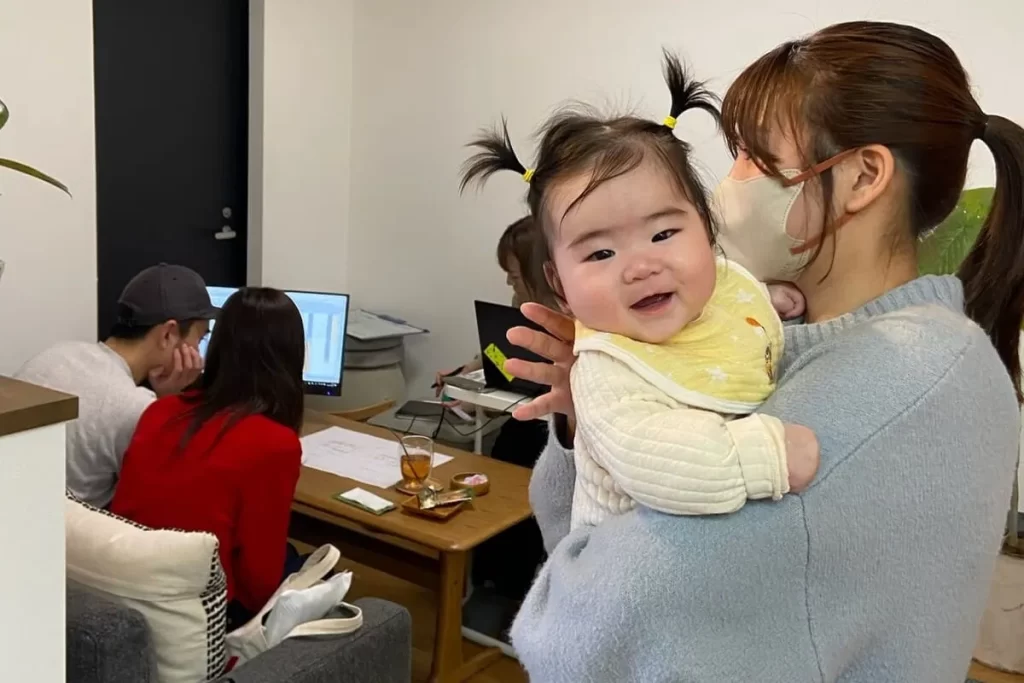 シンズクラフトの打ち合わせスペースで、赤ちゃんを抱っこするお母さんと、奥で資金計画の相談をするご家族の様子。