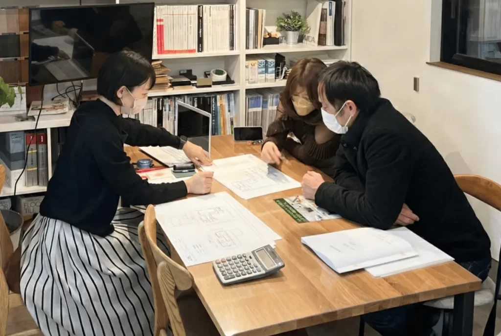 住宅会社のスタッフとテーブルを囲み、図面を見ながら打ち合わせをする夫婦の様子。