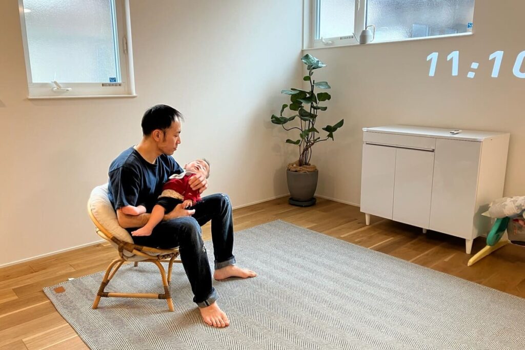 無垢床の明るいリビングで、赤ちゃんを抱っこしてくつろぐ男性の様子。