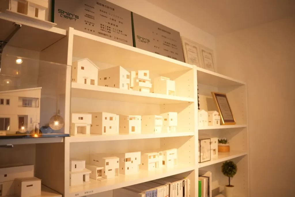白い棚に並べられた多数の住宅模型と、壁に掲げられたシンズクラフトの建設業許可証。