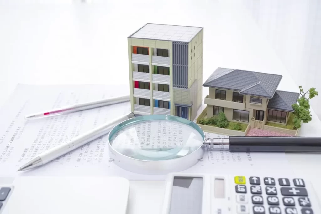 住宅模型、書類、虫眼鏡、電卓が並べられたデスクの上で、火災保険のシミュレーションを行っているイメージ。