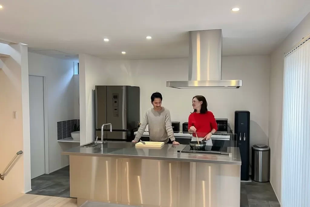 明るいアイランドキッチンのあるLDKで、笑顔で会話を楽しみながら料理をする若い夫婦の日常風景。